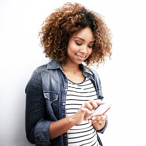 A smiling woman looking at her phone while leaning against a white wall