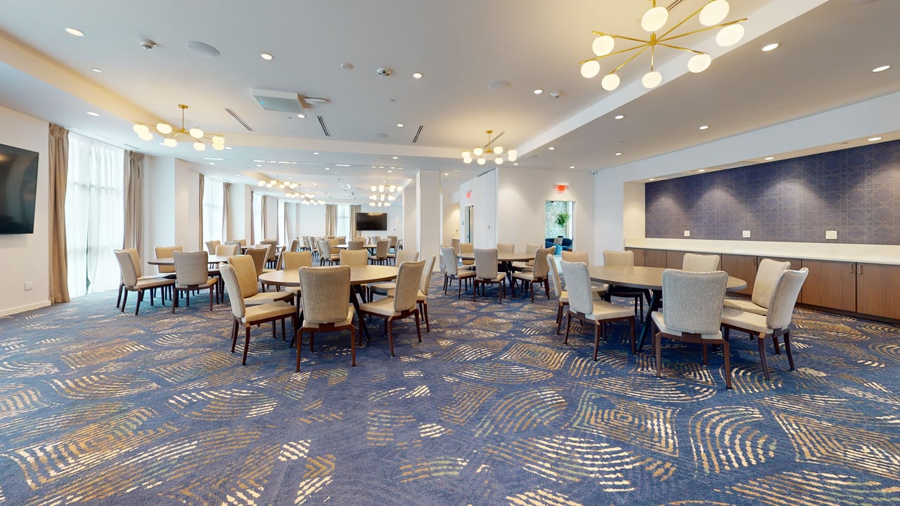 A hotel ballroom set with round banquet tables and cream chairs beneath gold chandeliers