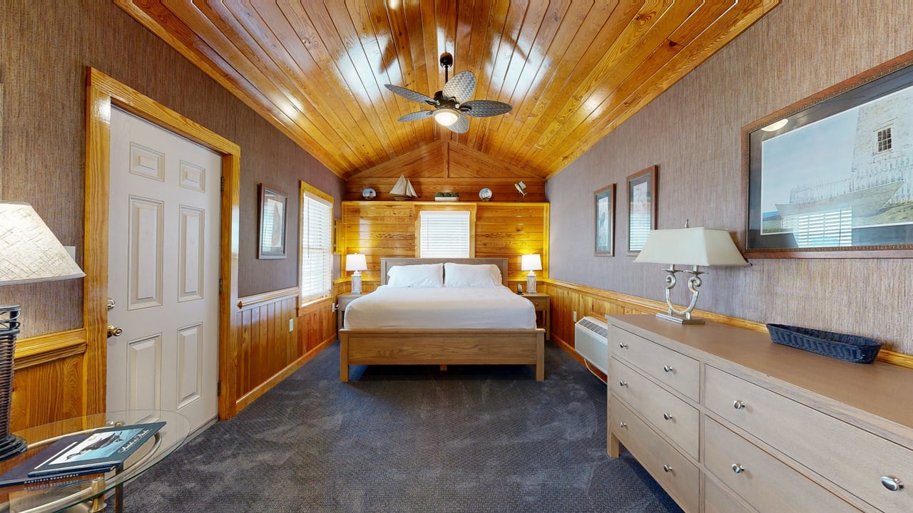 A cottage bedroom with a vaulted pine ceiling, king bed and framed nautical artwork