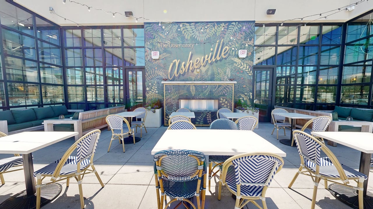 A glass-walled rooftop terrace with bistro tables, string lights and a painted botanical mural